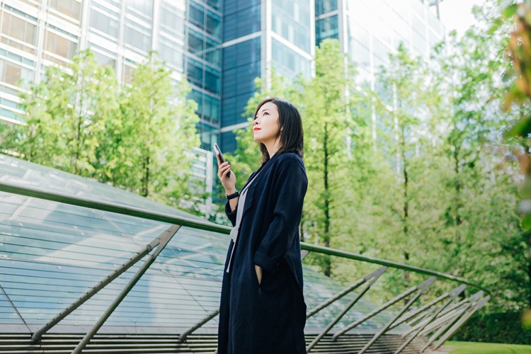 A young Asian woman holding a smart phone standing against corporate buildings