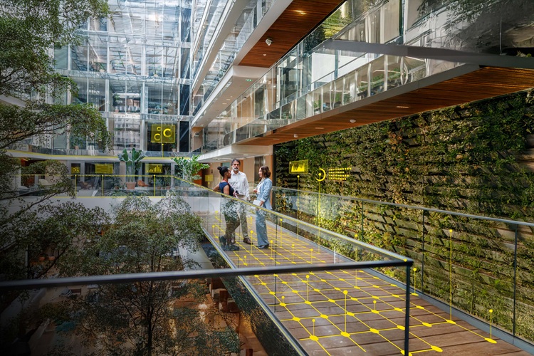 Three business professionals of diverse backgrounds engage in a discussion while standing in a spacious, well-lit office hallway featuring modern architecture and greenery.
