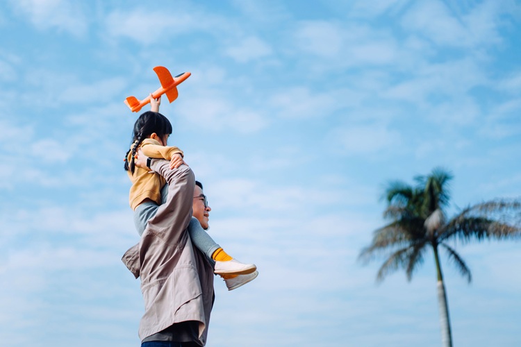 Joyful young Asian father carrying his little daughter on shoulders, spending time together outdoors, playing with airplane toy in park on a lovely sunny day against beautiful blue sky